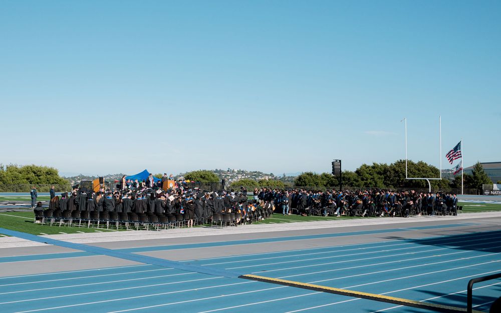 Military graduates in formation stand on a blue field during an outdoor graduation ceremony under clear blue skies.