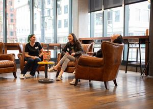 Two people having an informal meeting while sitting in leather armchairs in a modern office space with large windows.
