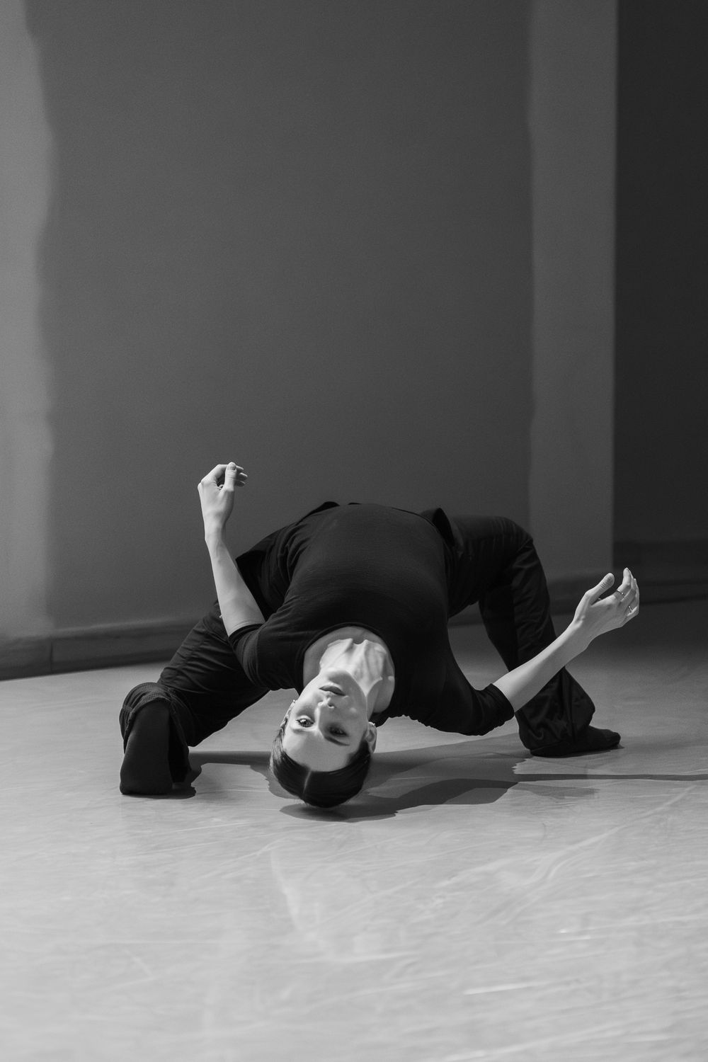 A dancer in black clothing performs a fluid contemporary dance movement on a light floor against a neutral background.