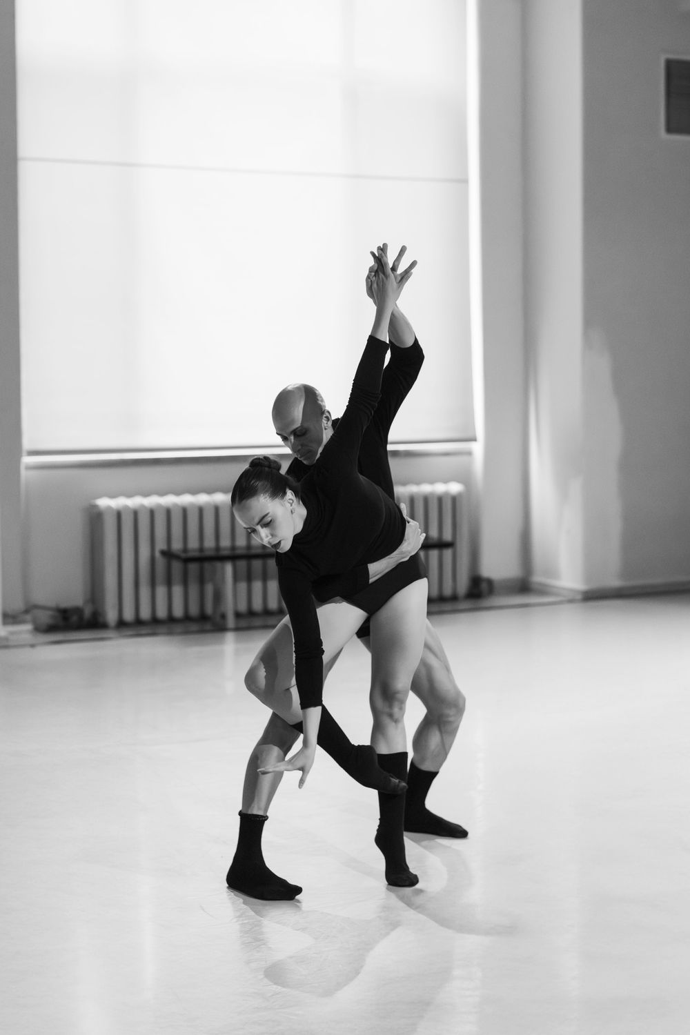 A dancer in a black leotard performs a dynamic modern dance pose with graceful arm extension in a bright studio.