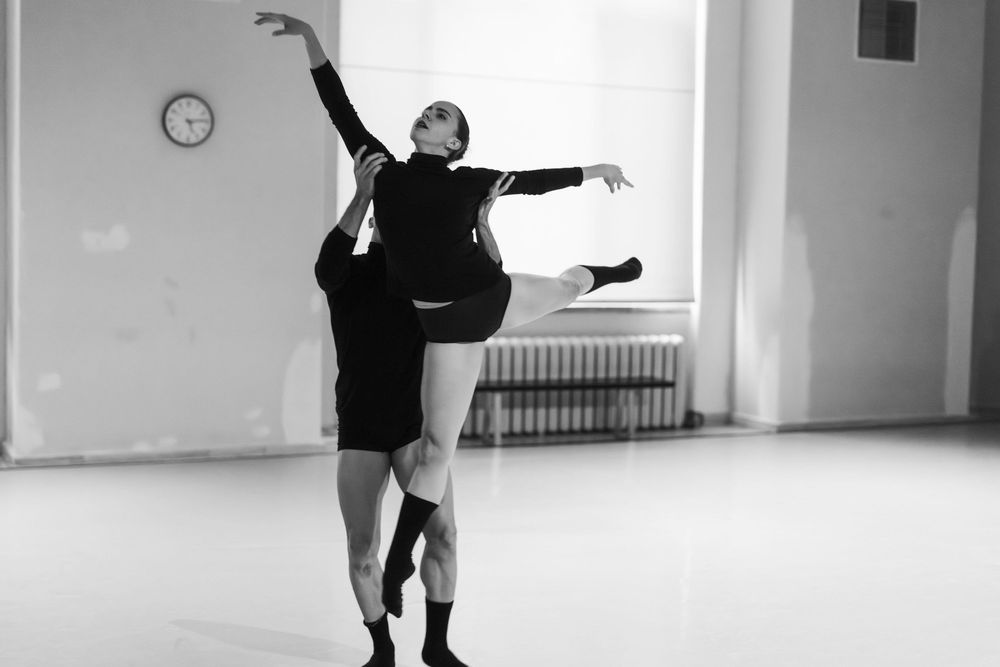 A dancer in black attire performs a graceful modern dance movement in a bright studio space.