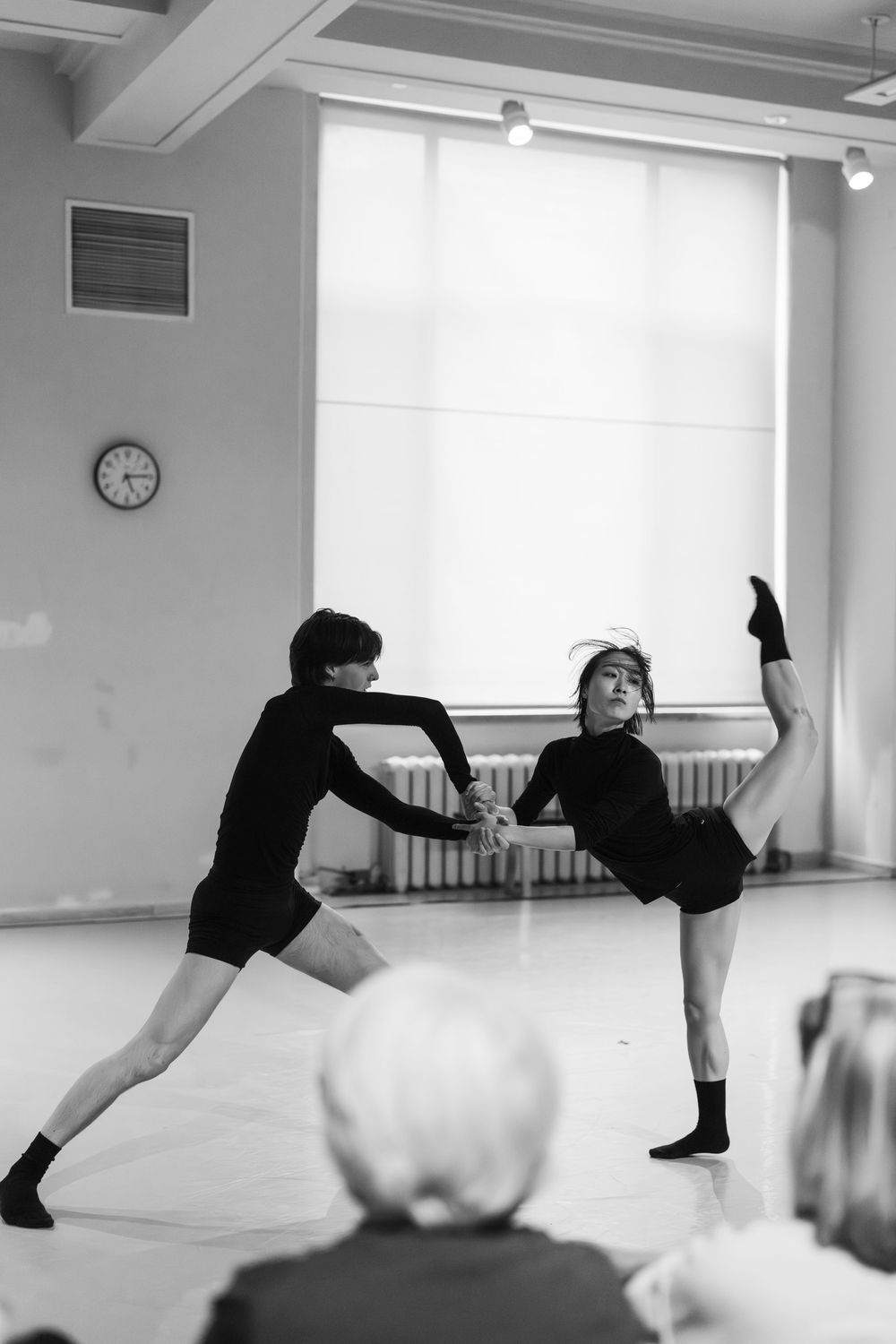 Two dancers performing a dramatic duet with high leg extensions in a bright studio while others watch.