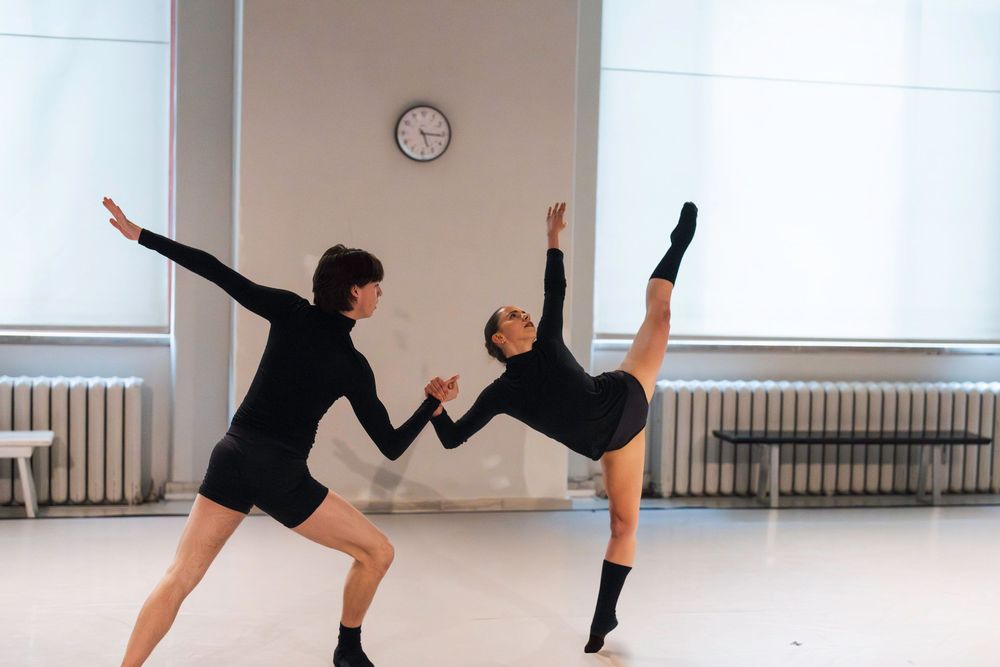 Two modern dancers in black outfits practice dynamic movements in a bright dance studio with clock on wall.