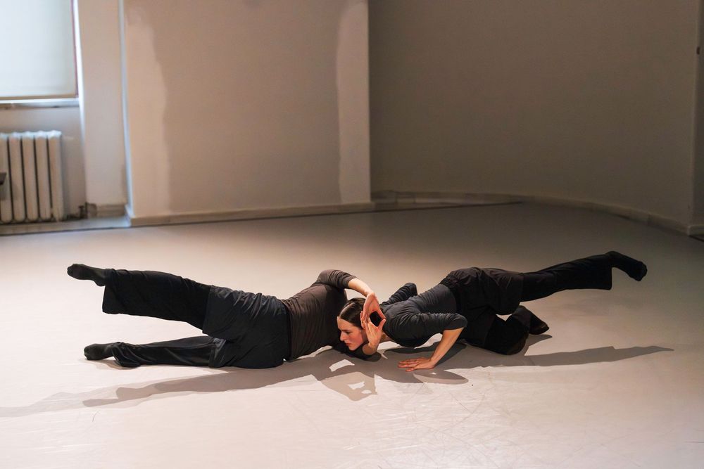 Two dancers in black outfits performing a dynamic floor routine in an empty studio space.