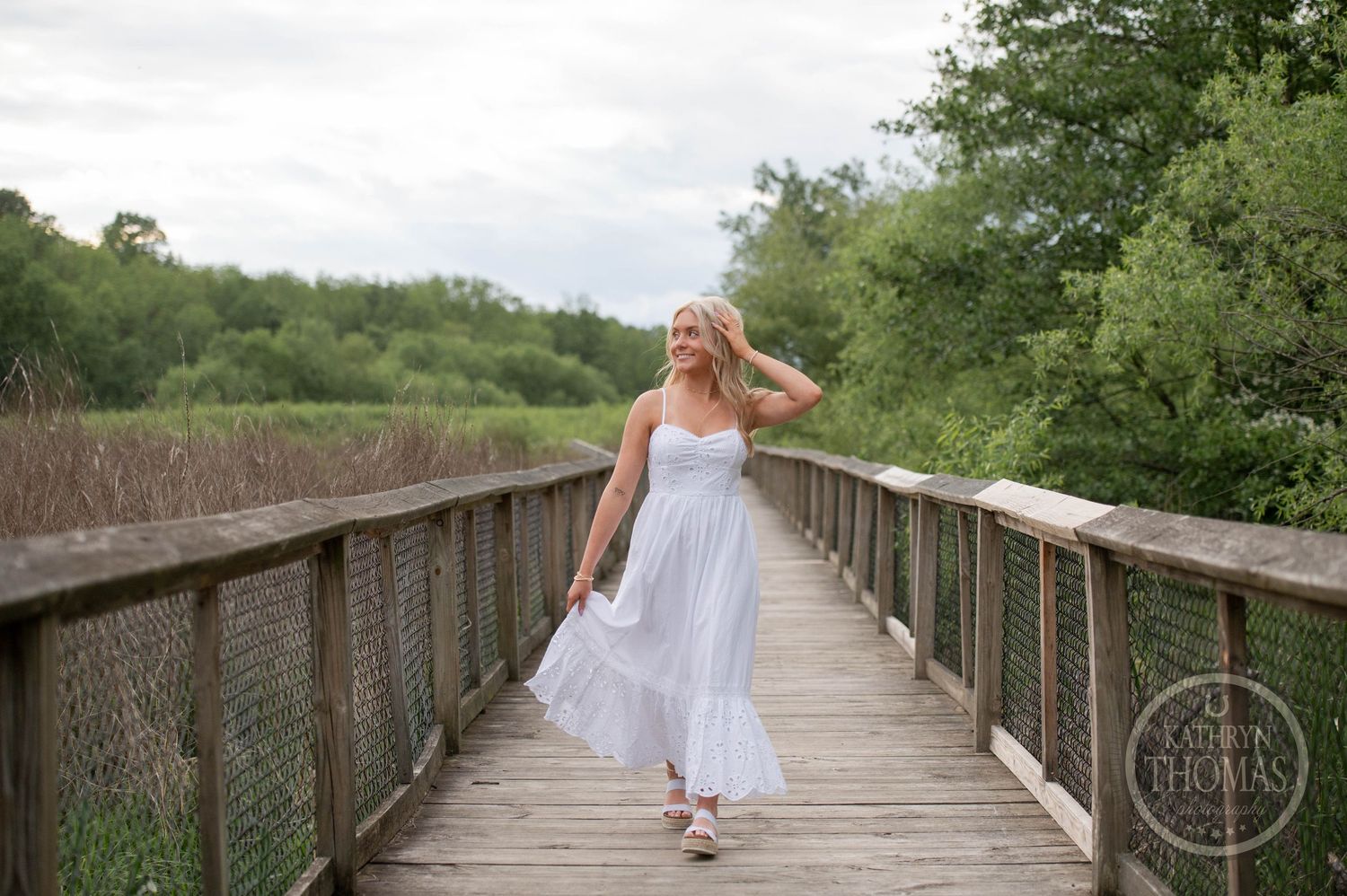 A person in a flowing white dress twirls on a wooden boardwalk surrounded by lush green vegetation.