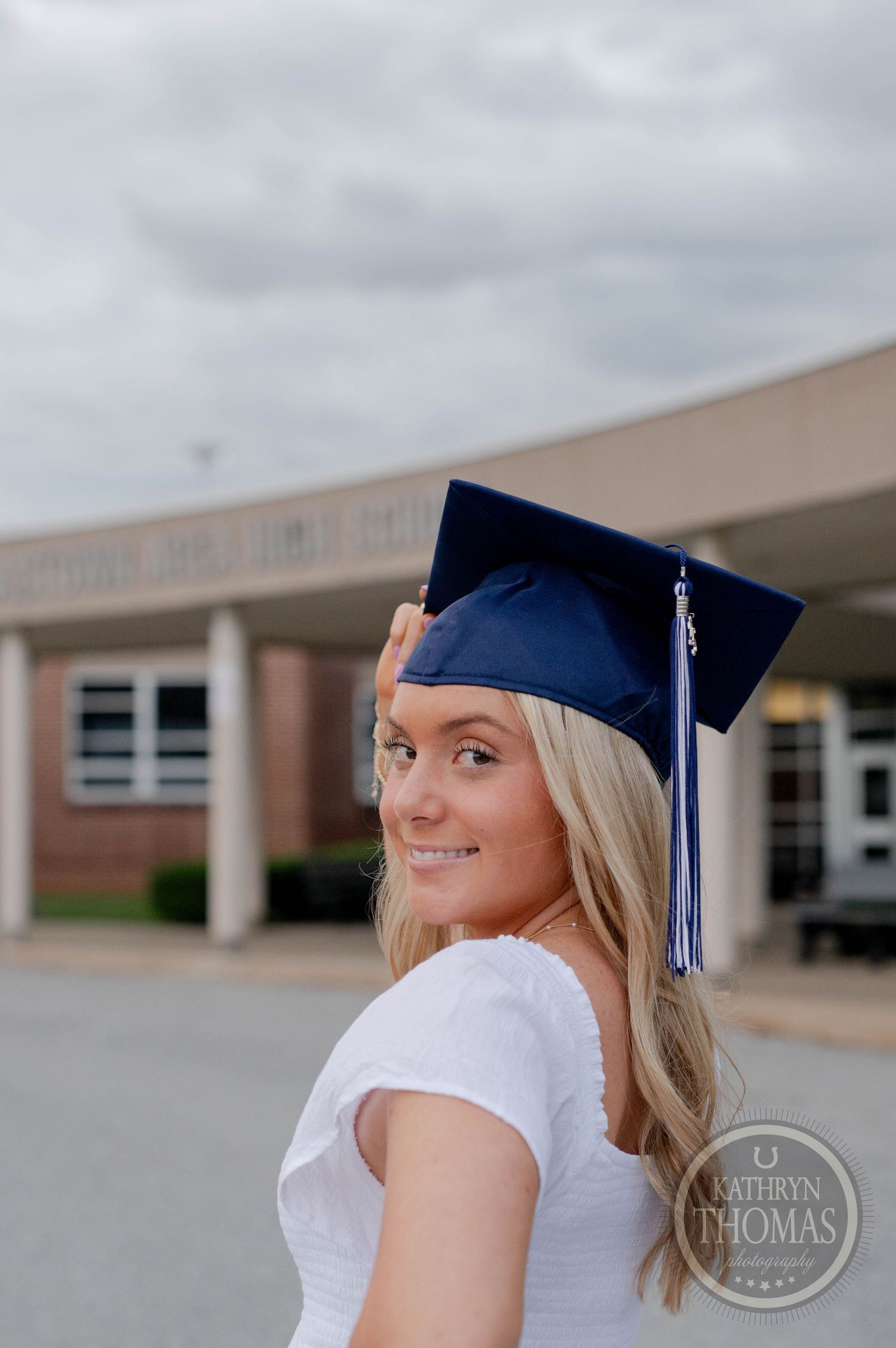 A graduate in a white top and blue cap with tassel poses for photos against a brick school building background.