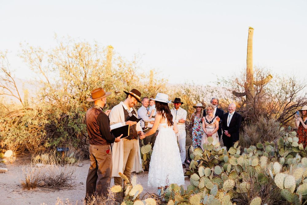 A small wedding gathering stands together in a desert landscape with cacti and golden hour sunlight.