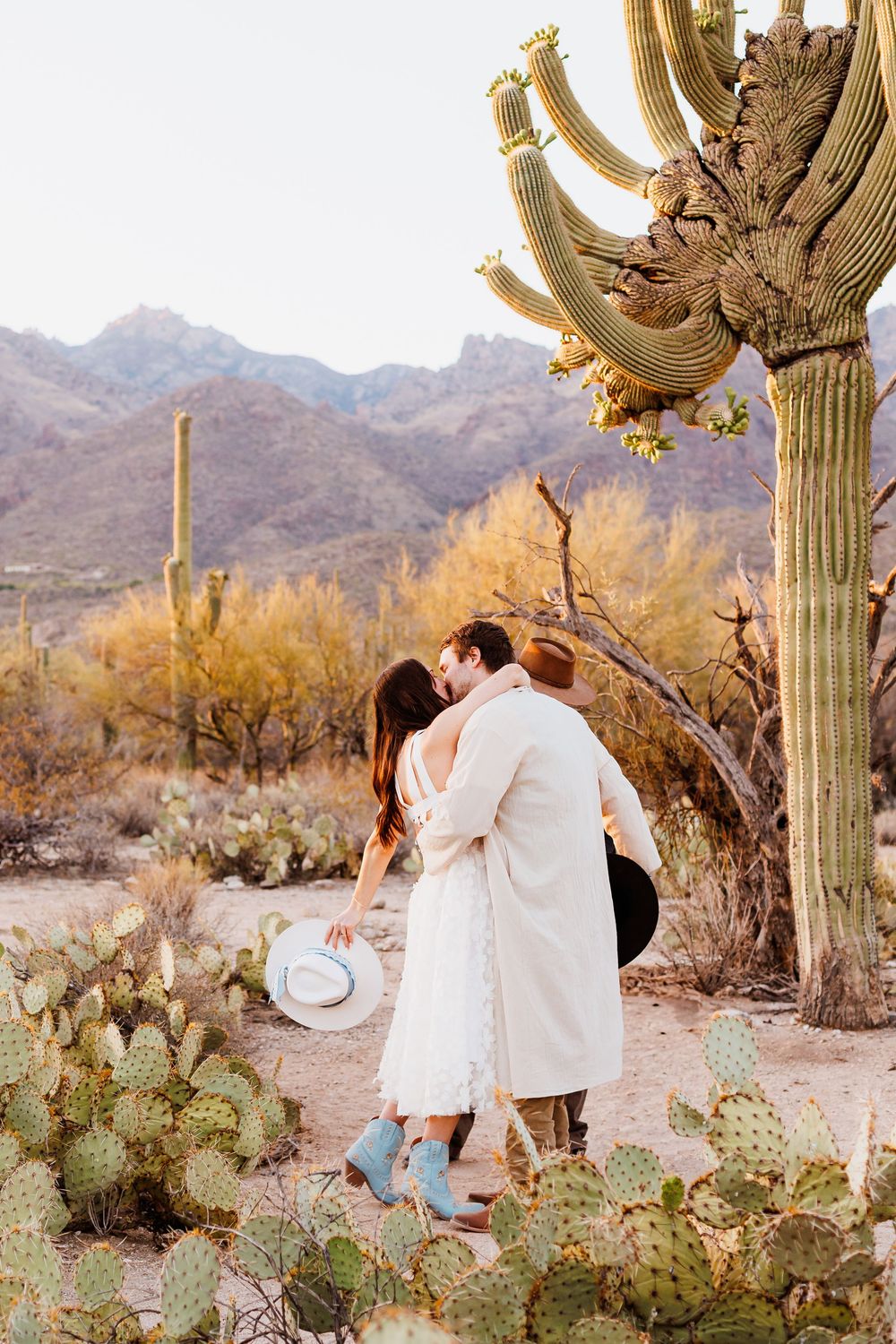 A couple embraces in a scenic desert landscape surrounded by saguaro cacti and mountains at sunset.
