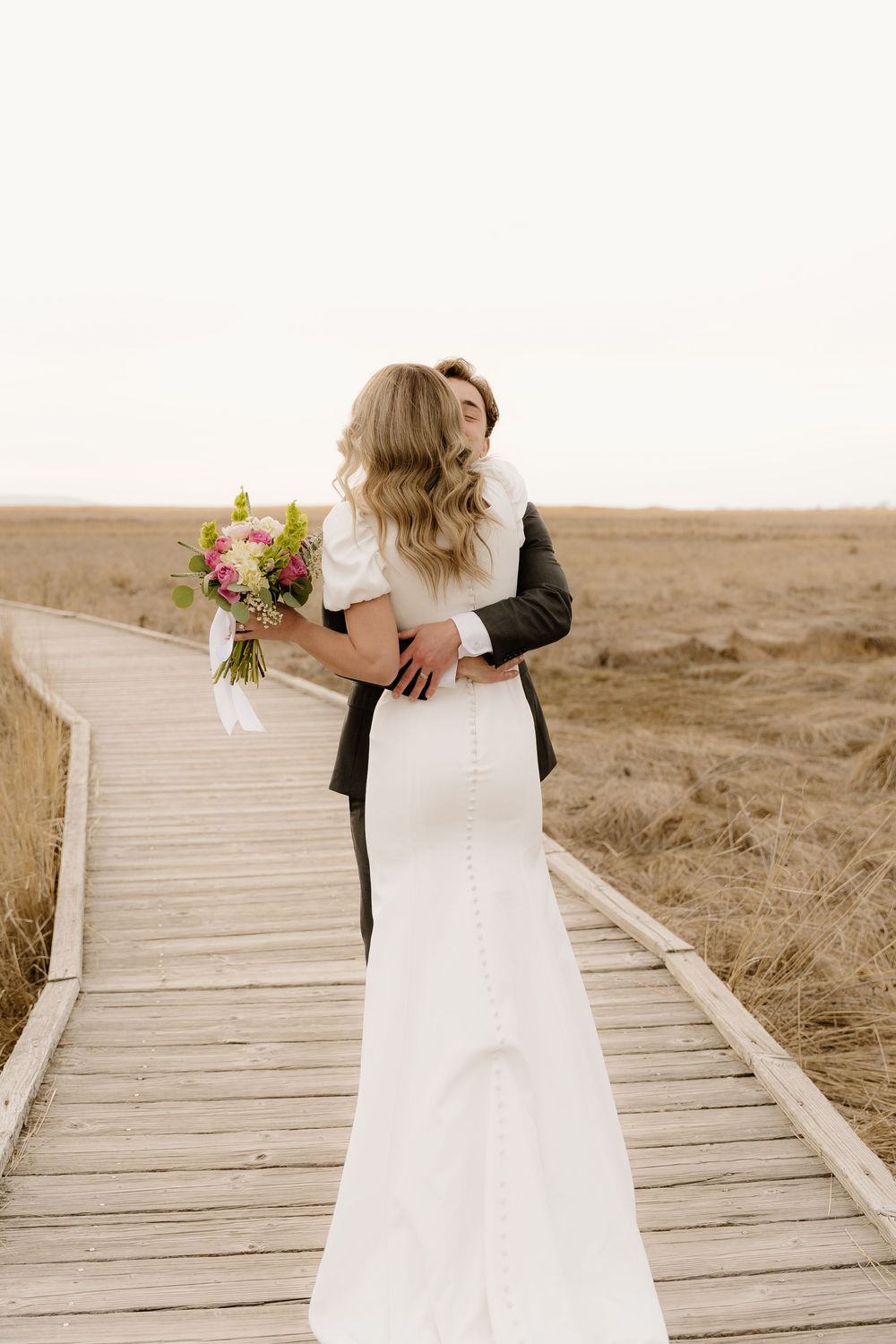 A romantic wedding couple embraces on a wooden boardwalk surrounded by dry grass at sunset.