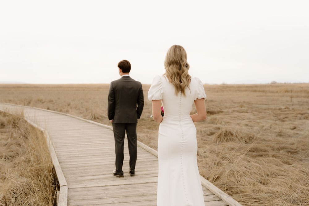 Bride and groom walk away together on a wooden boardwalk stretching through golden prairie fields.