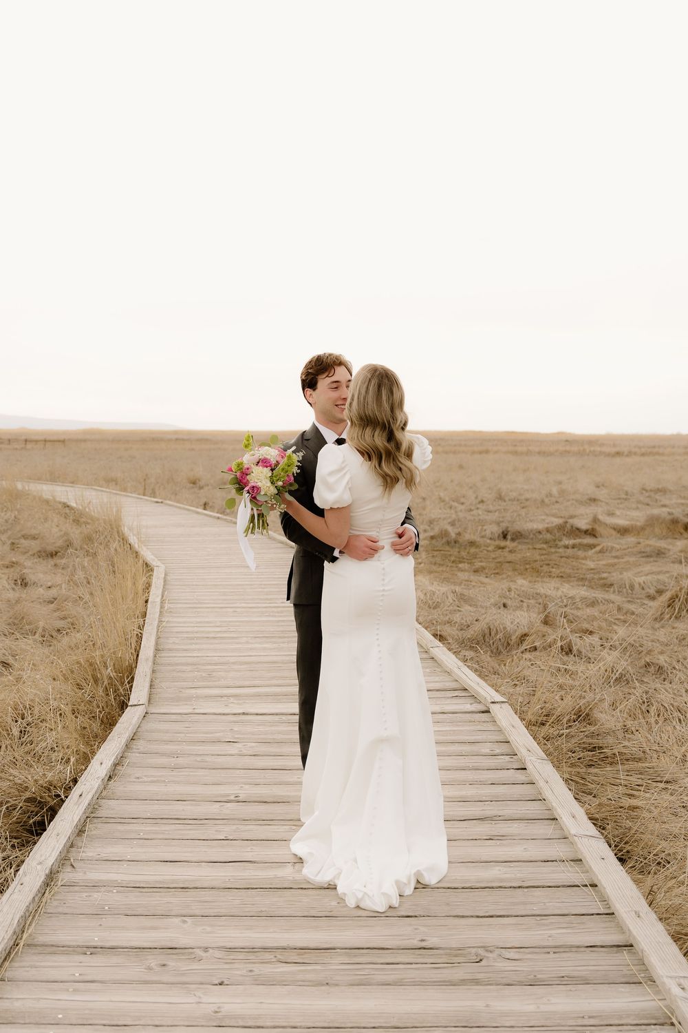 Romantic wedding couple embracing on wooden boardwalk surrounded by golden prairie grasses.