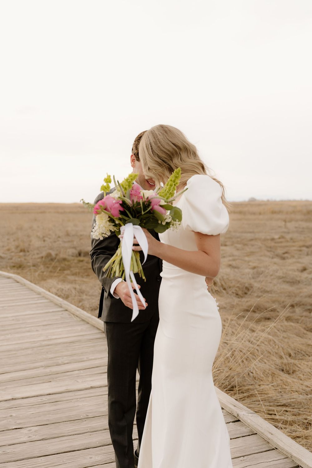 Romantic wedding photo of couple embracing on wooden walkway with bride holding floral bouquet.