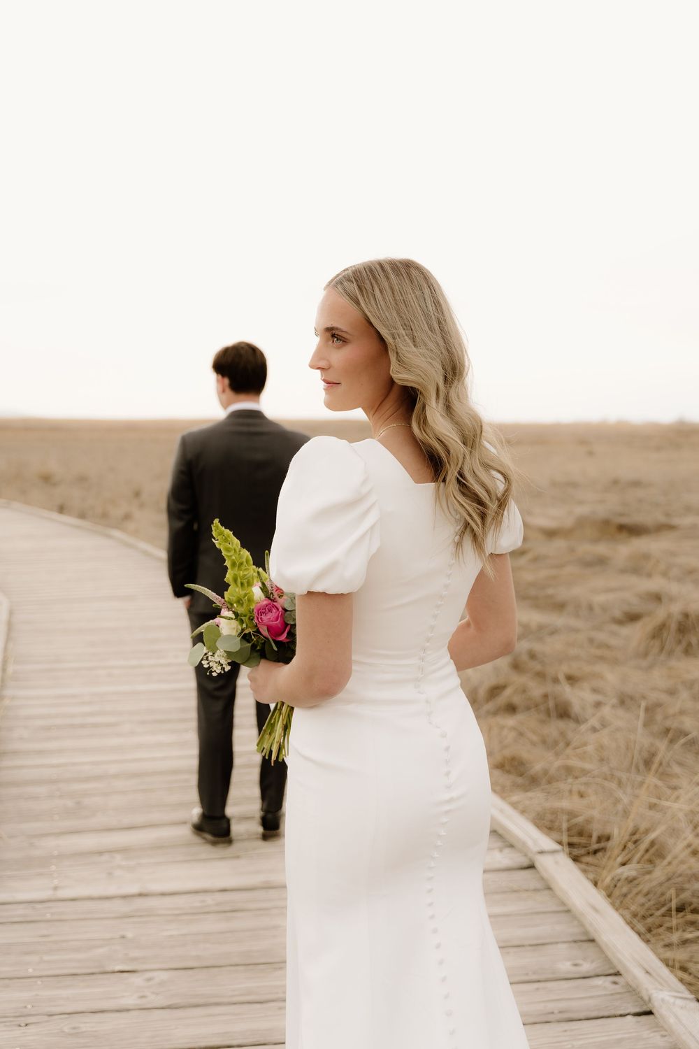 Bride in fitted white dress holding bouquet while groom stands in distance on wooden boardwalk through marsh.