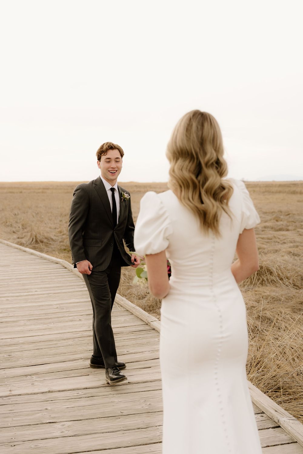 Bride in white dress and groom in dark suit stand facing each other on a wooden boardwalk in a rustic field.
