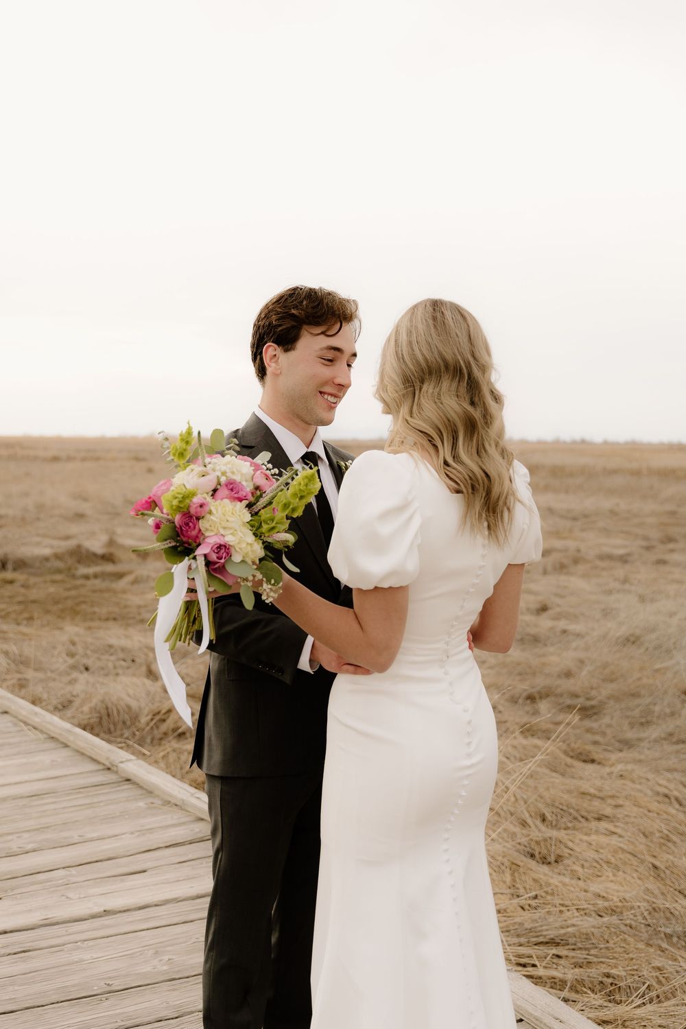 Wedding couple shares an intimate moment on a boardwalk path through a desert landscape with colorful bouquet.