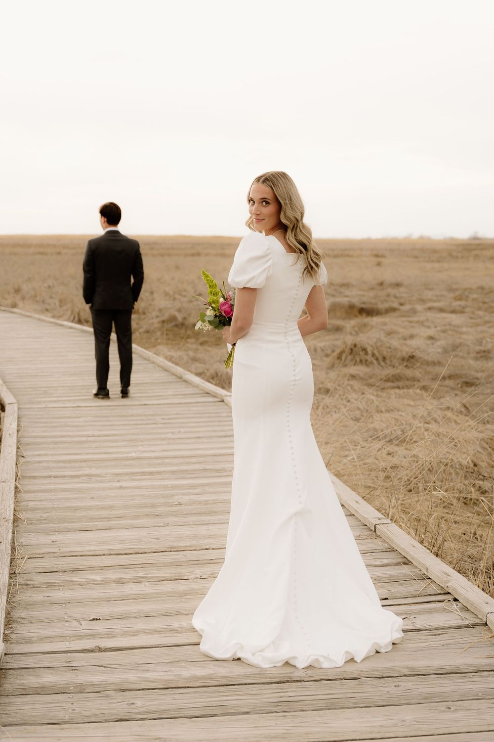 Bride in elegant fitted white gown stands on wooden boardwalk with scenic prairie landscape in background.