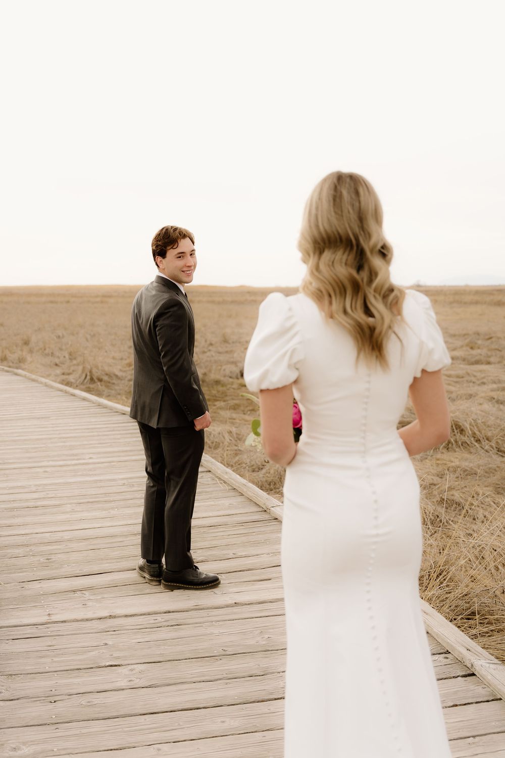 A couple in wedding attire stand apart on a wooden boardwalk with golden prairie grass in the background.