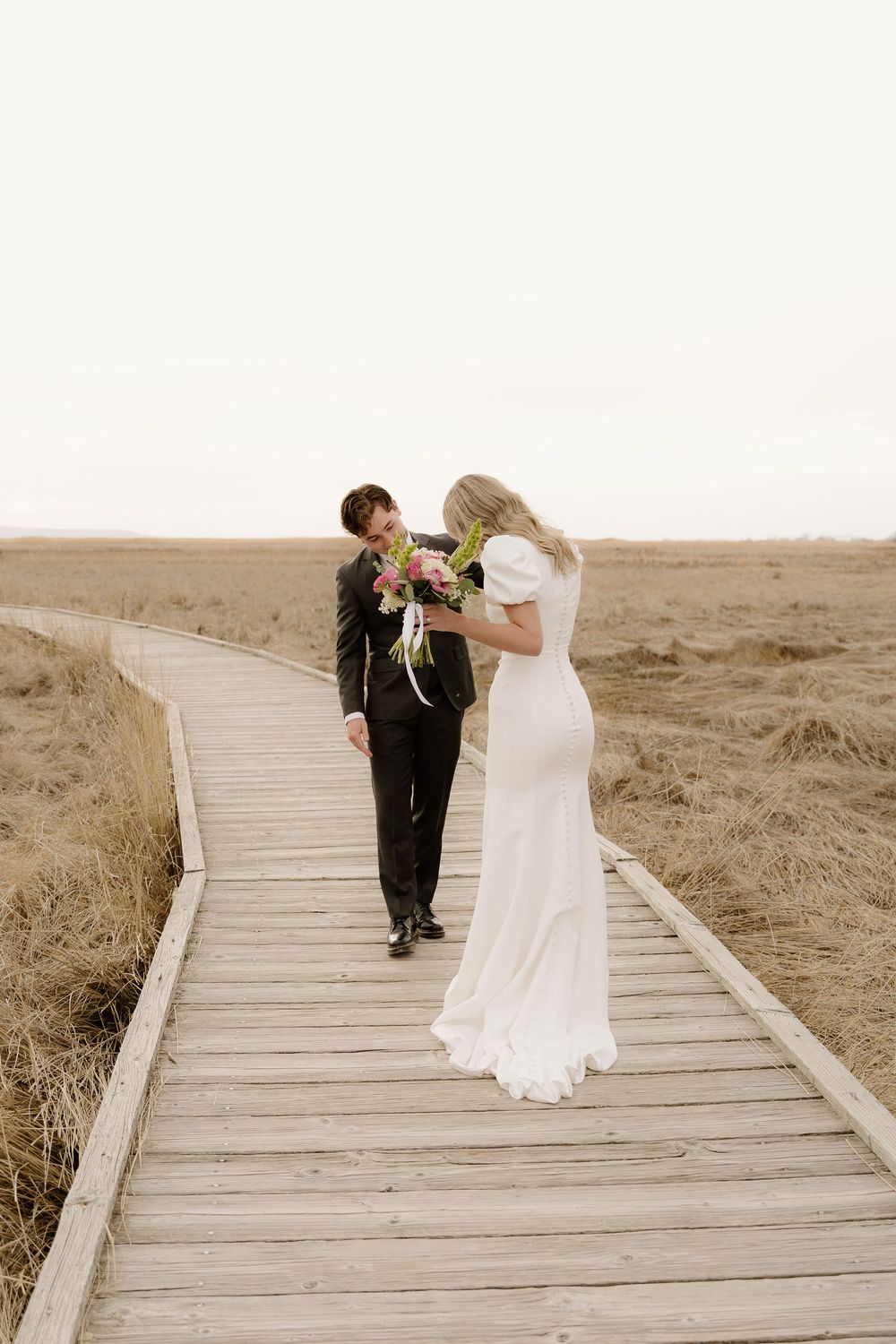 Newlyweds share an intimate moment on a wooden boardwalk surrounded by golden grass and soft light.