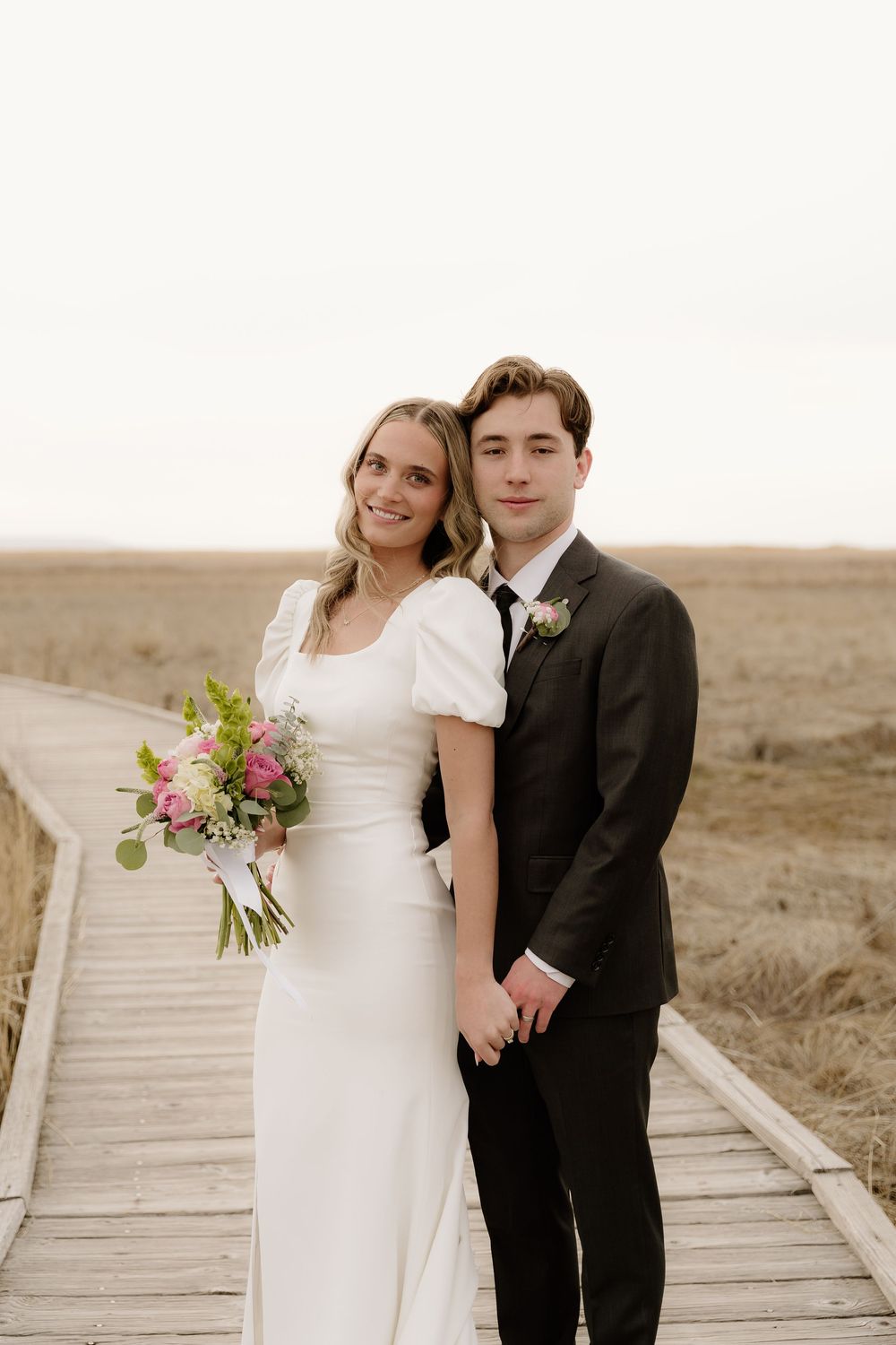 Newlyweds pose together on wooden boardwalk surrounded by golden fields.
