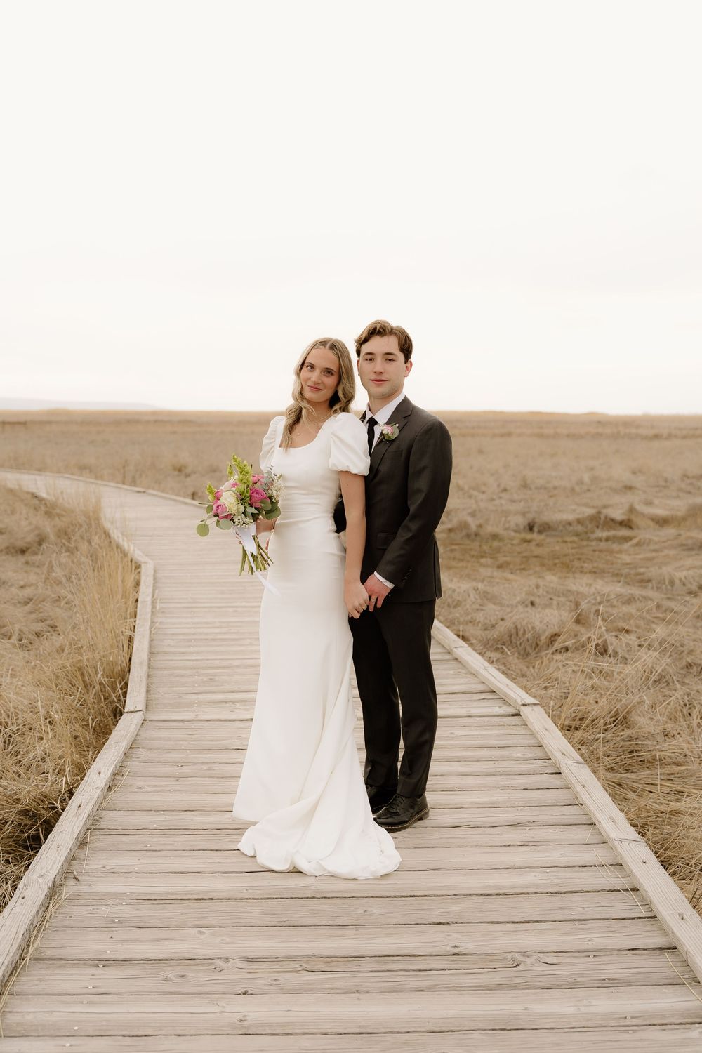 Wedding couple stands embracing on wooden pathway through serene marshland landscape.