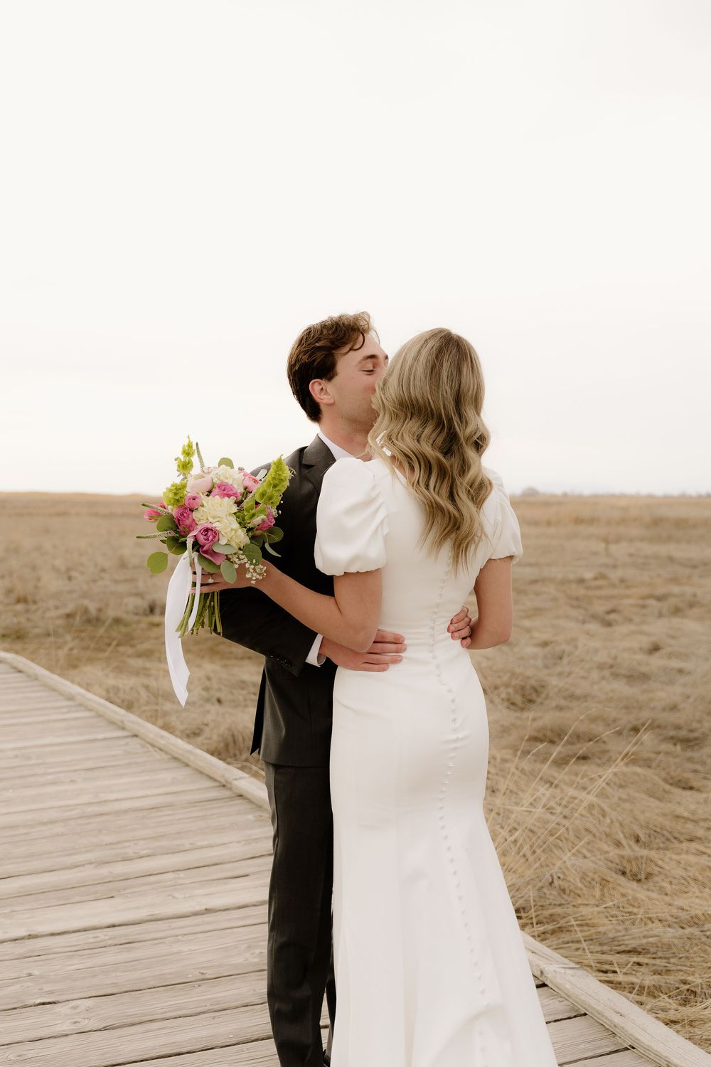 A couple embraces on a wooden boardwalk surrounded by dry grass field while holding a pink and white floral bouquet.