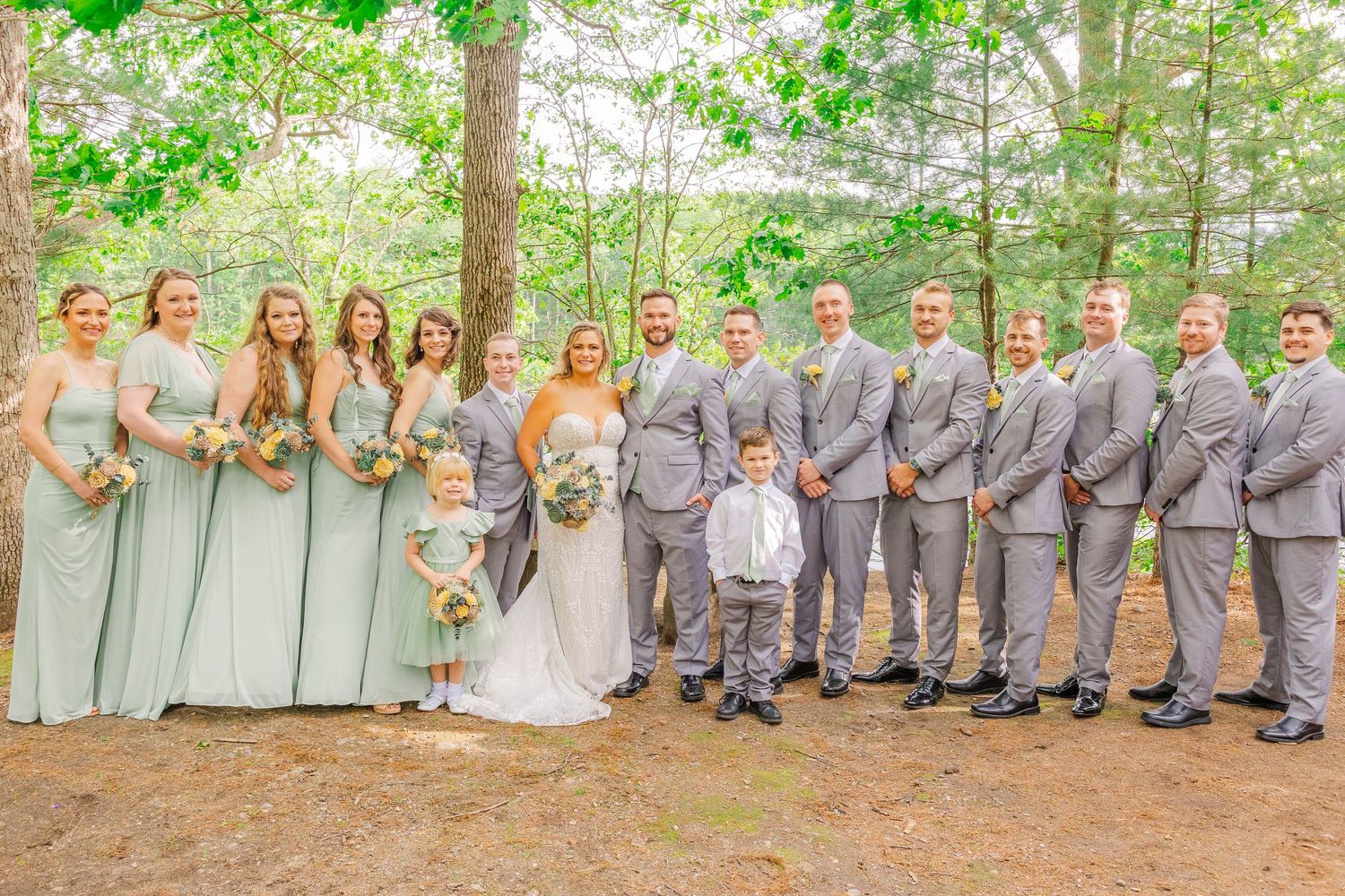Wedding party in sage green and gray attire poses together in a wooded outdoor setting during celebration.