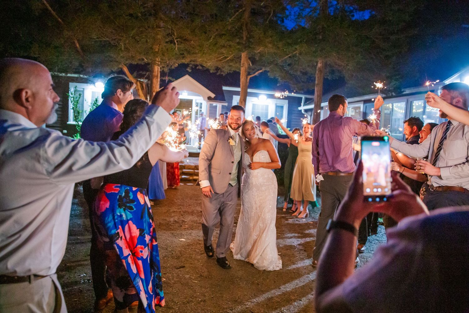 Wedding guests dance and celebrate with sparklers during an outdoor evening reception at a wooded venue.