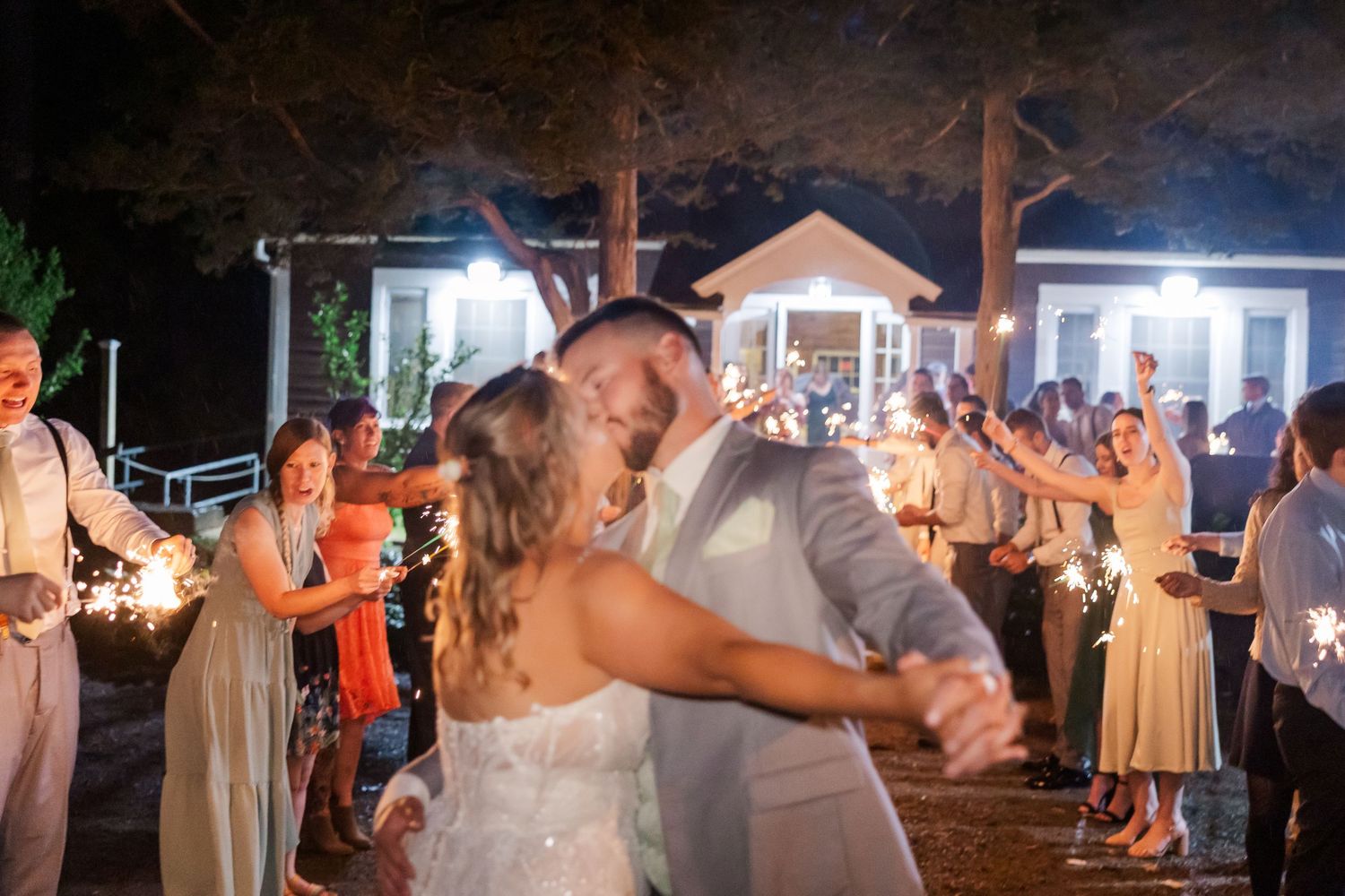 Newlyweds share their first dance surrounded by guests holding sparklers during an evening outdoor wedding reception.