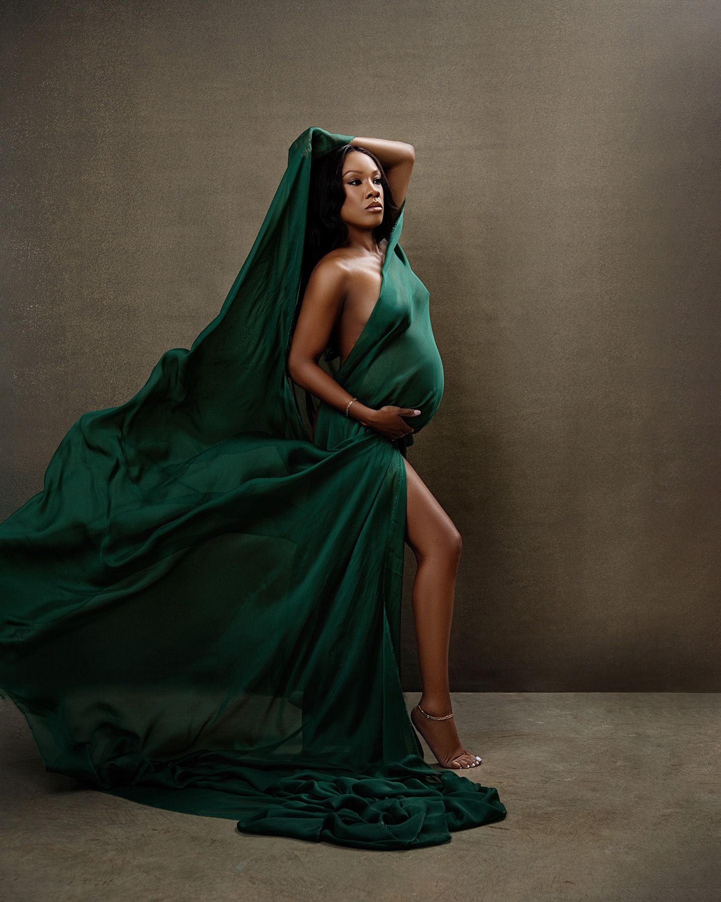 A figure in an emerald green flowing gown poses dramatically against a neutral backdrop with elegant lighting.