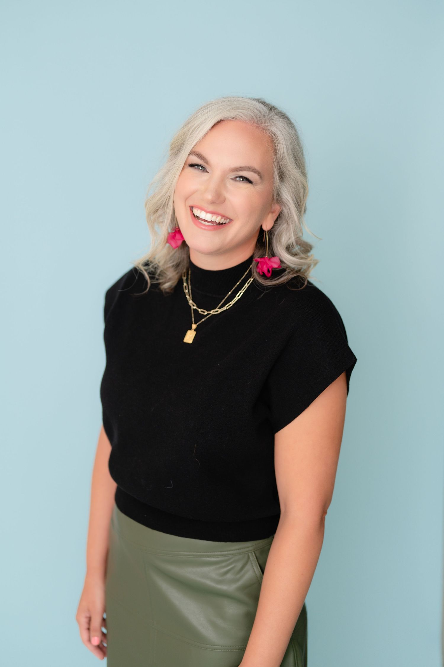 Professional portrait of someone wearing a black top and olive skirt against a light blue background, with statement jewelry.