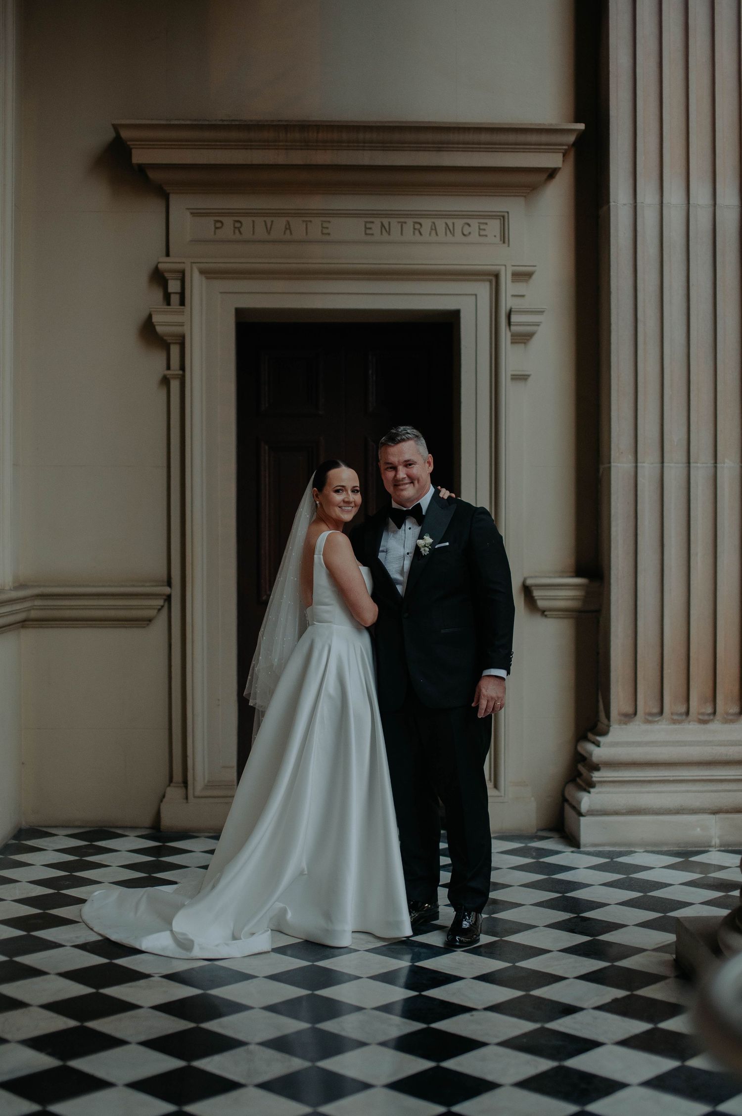 A newlywed couple poses in front of a private entrance with a classic black and white checkerboard floor.