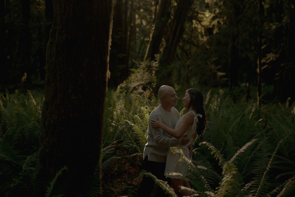 A couple embraces in a forest clearing surrounded by tall ferns and trees.