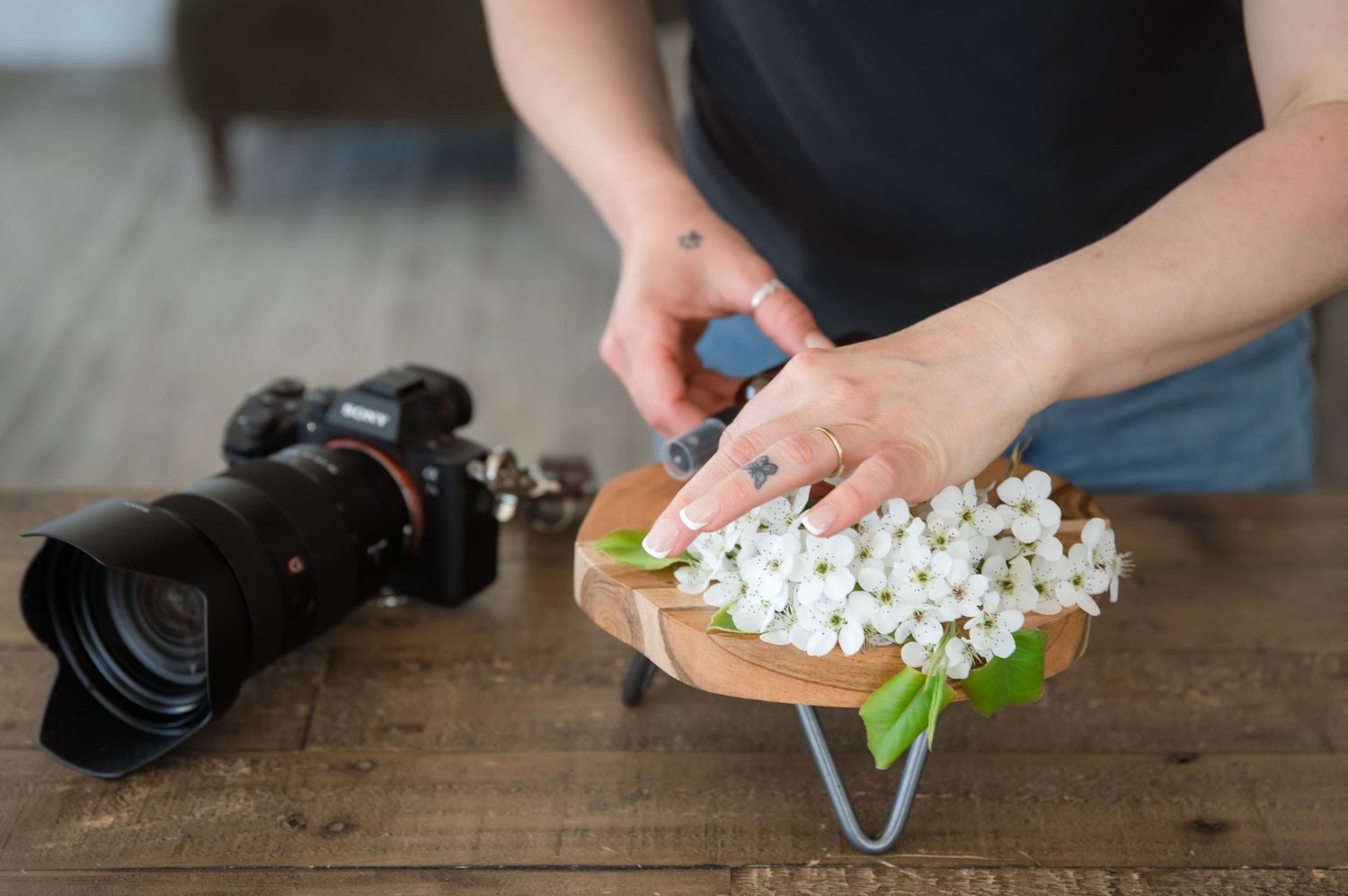 Someone arranging small white flowers on a wooden table next to a black camera for a photography session.