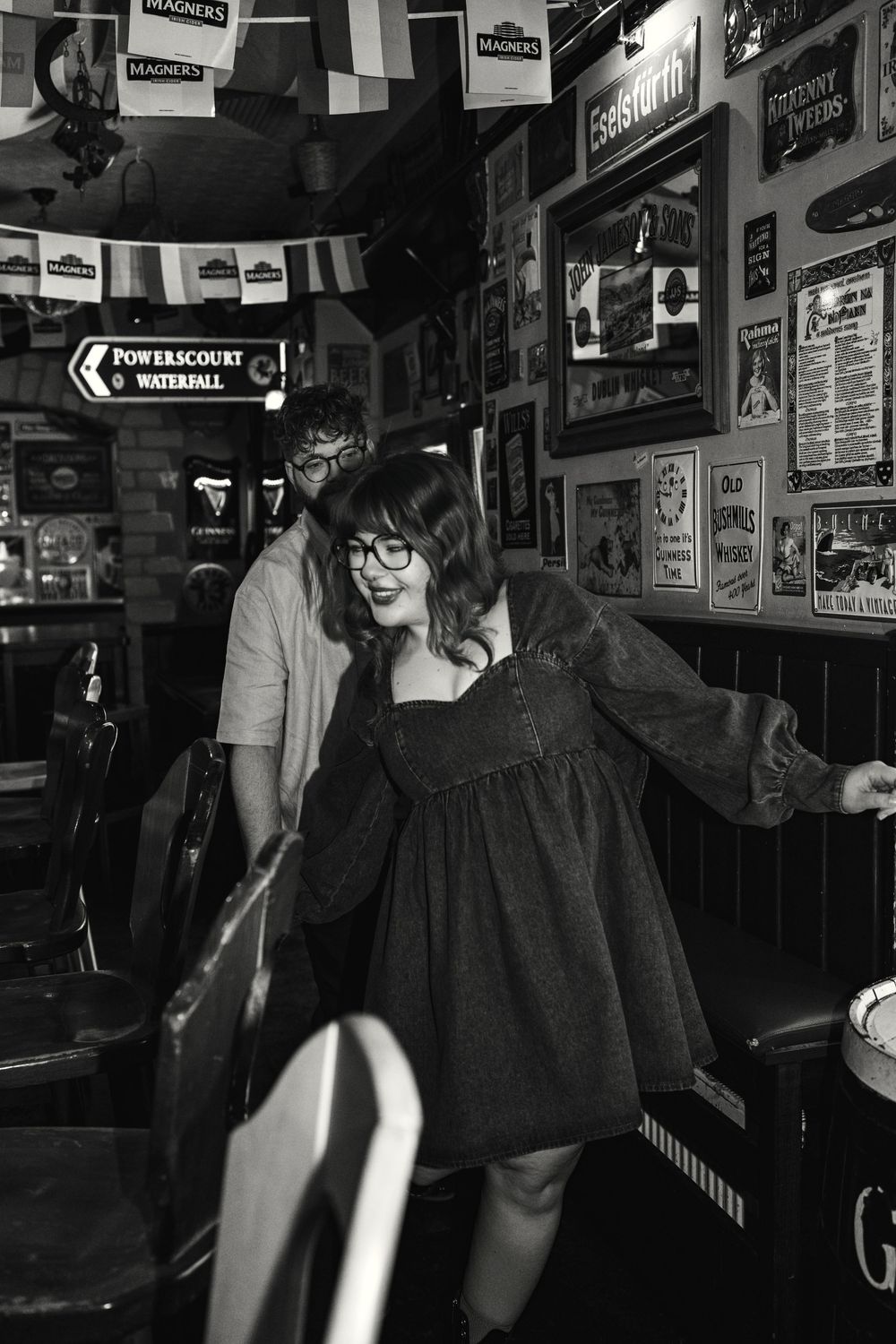 Black and white photo shows person in retro dress and glasses dancing playfully inside a dimly lit pub with posters on walls.
