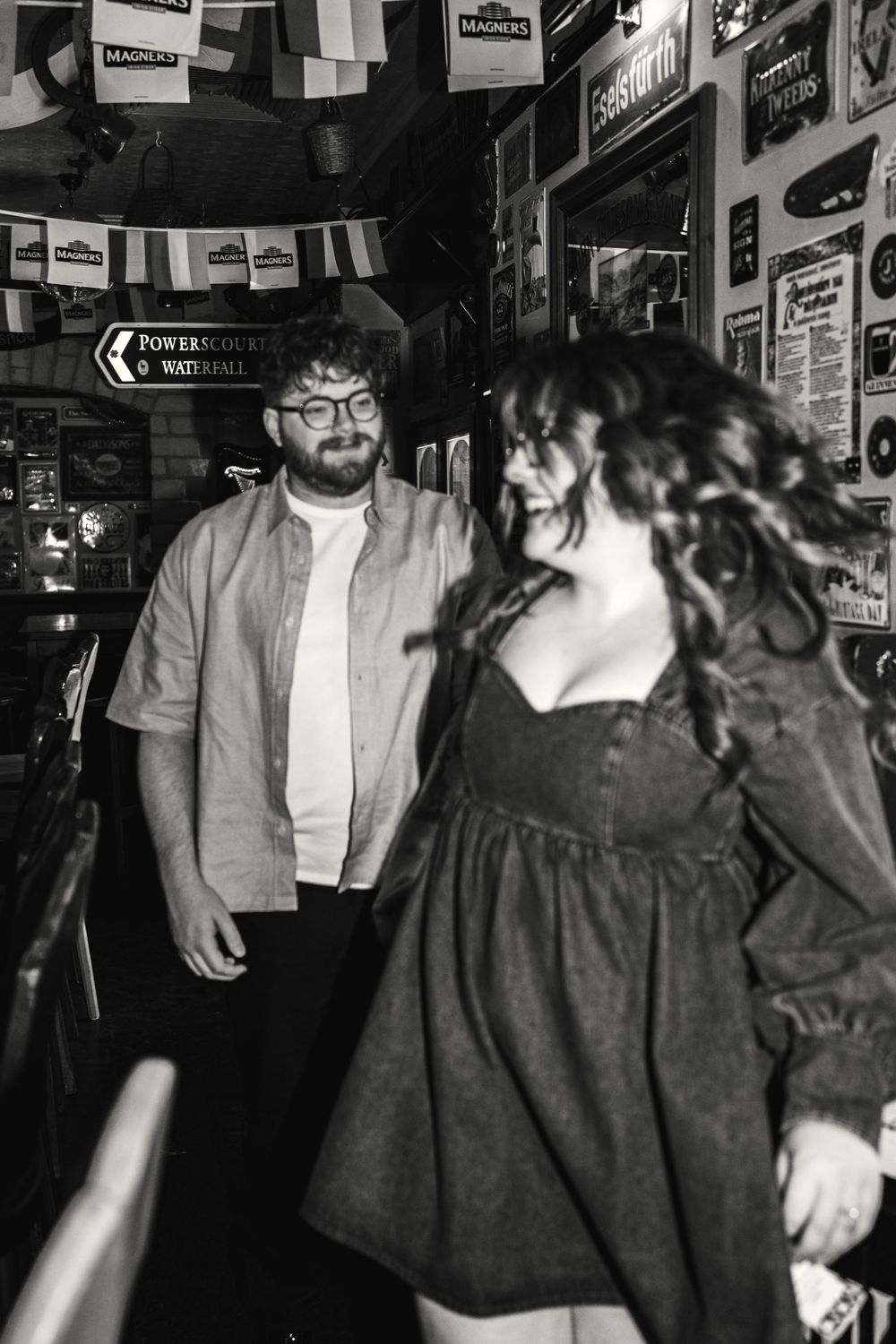 Black and white photo of two people laughing and socializing inside a bar decorated with hanging banners and posters.
