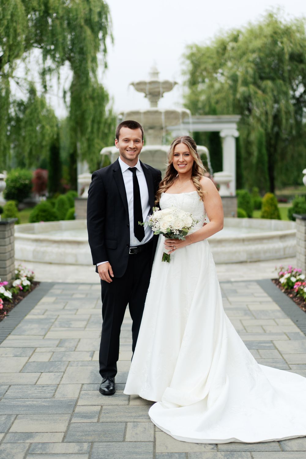 Wedding couple stands on stone path in front of fountain and willow trees in elegant garden setting.