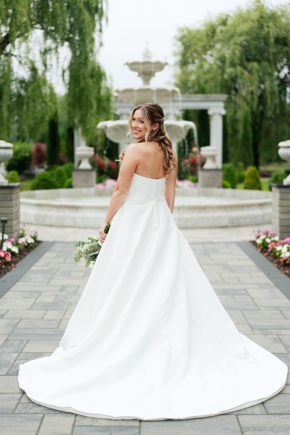 Elegant white wedding gown with long train photographed from behind in a garden setting with fountain and willow trees.