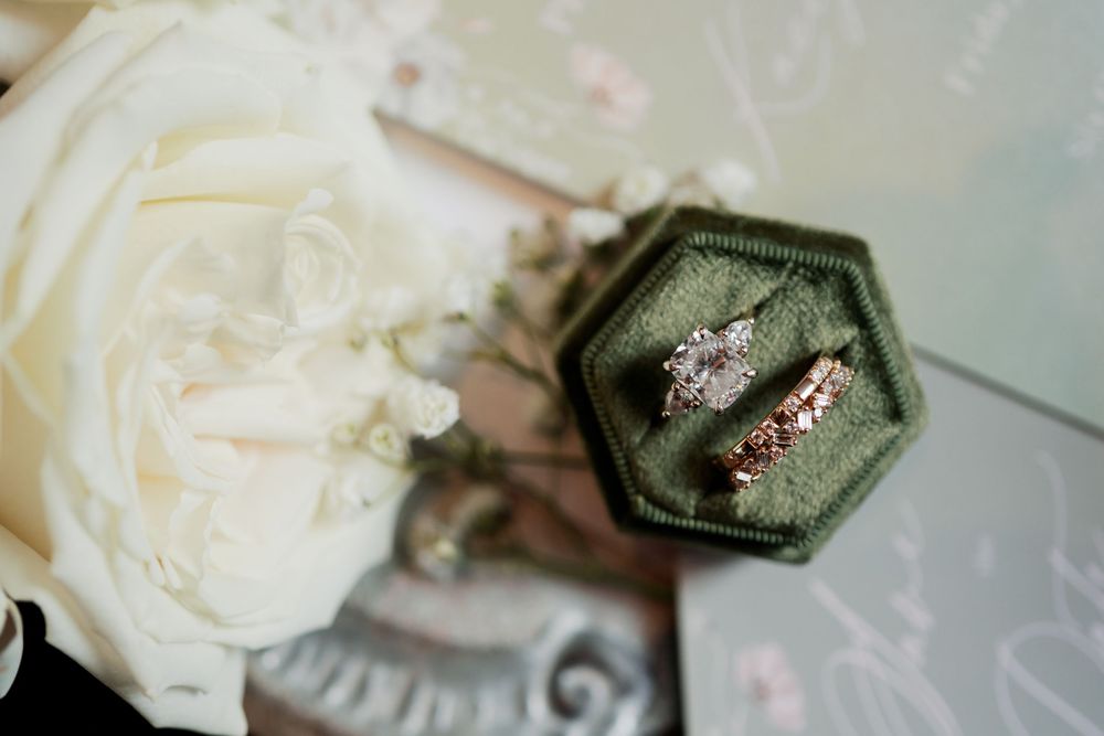 Two diamond engagement rings rest in a dark green velvet hexagonal ring box beside a white rose.