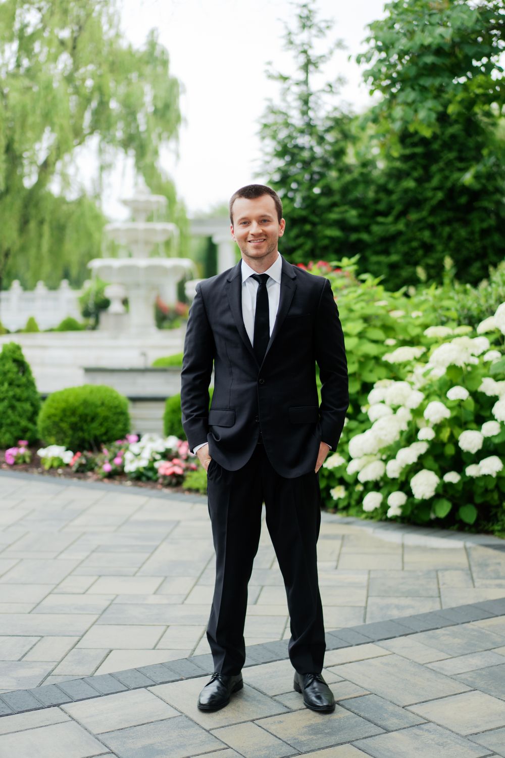 A person in a black suit and tie stands on stone pavers in a manicured garden with white flowers and a fountain.