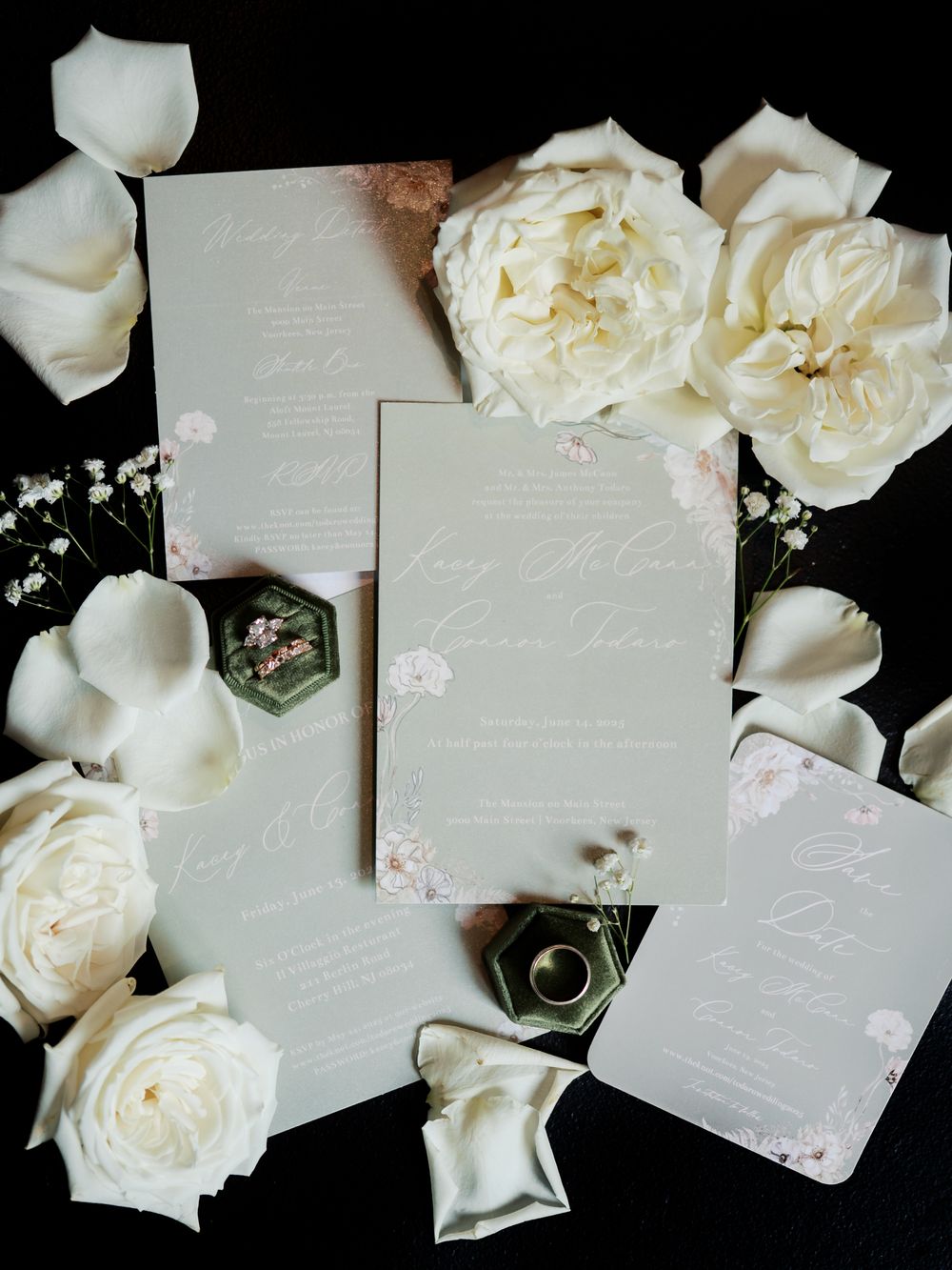 Elegant white wedding invitations and roses arranged on black background with delicate floral details and rings.