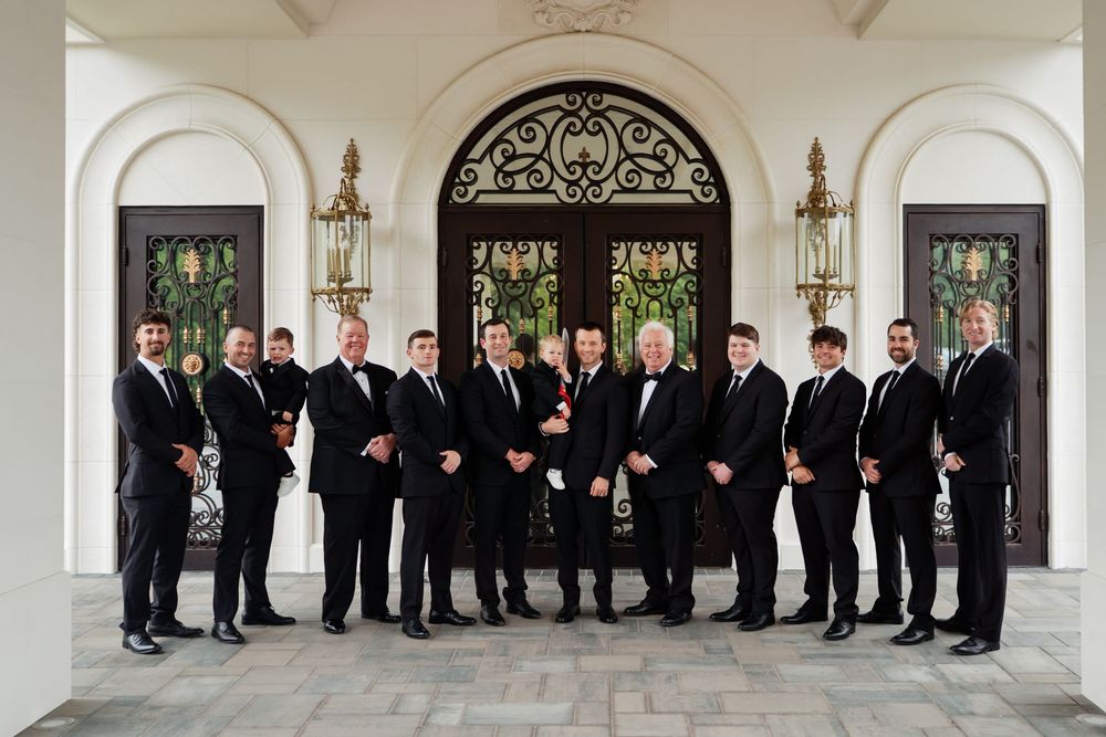 A group in formal black suits and ties stands together in front of an elegant archway entrance with lanterns.