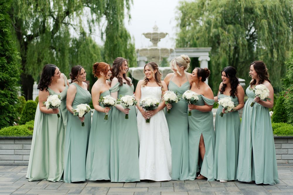 Wedding party in sage green dresses pose together in front of weeping willow trees in a garden setting.