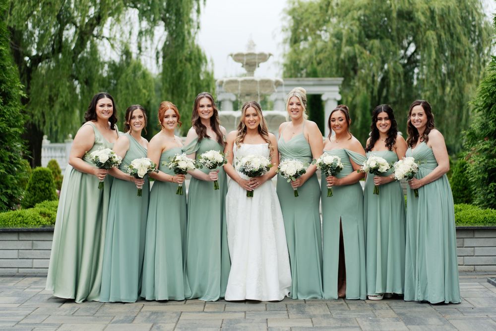 Wedding party poses in front of fountain wearing matching sage green dresses and holding floral bouquets.