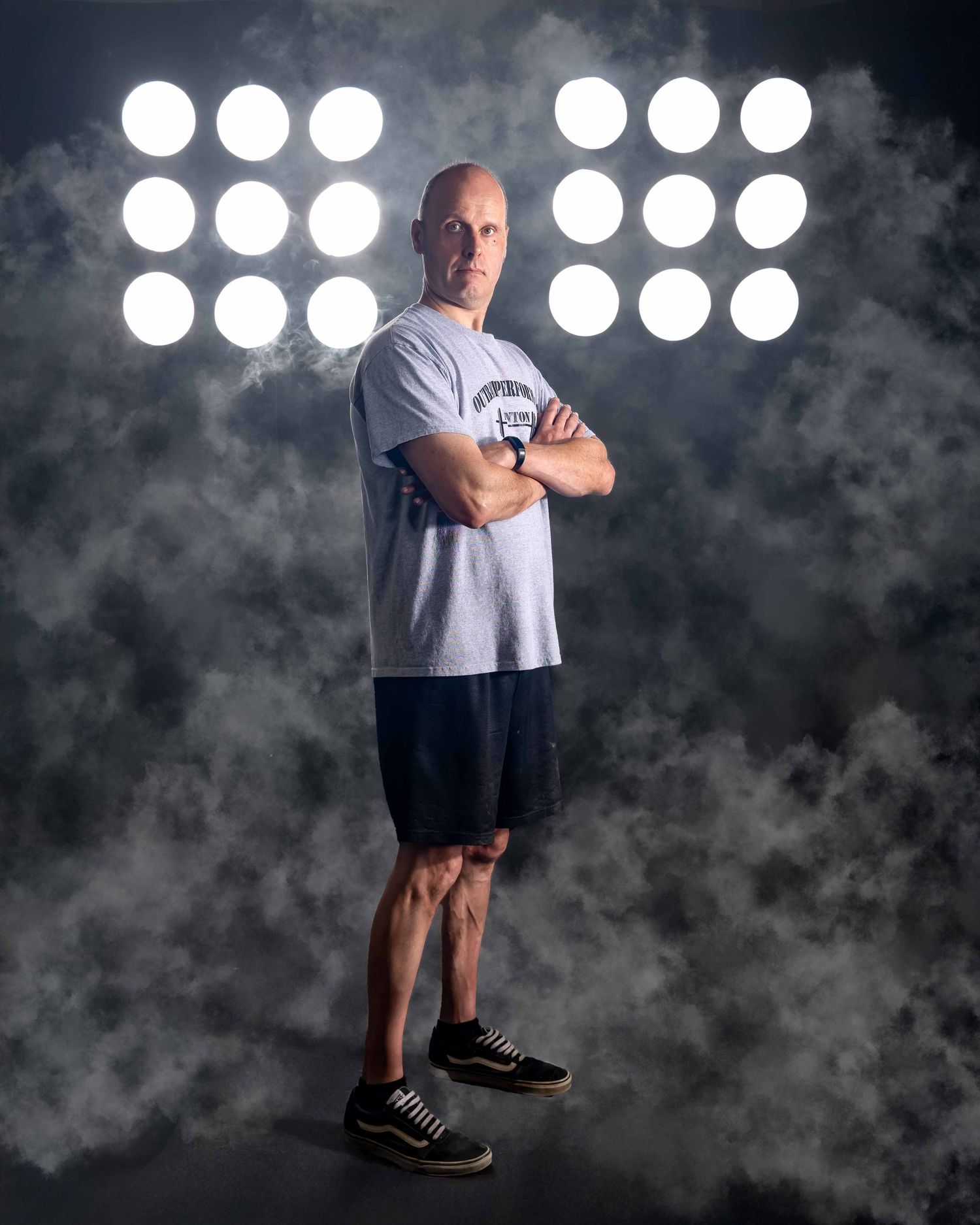 Person in white t-shirt stands with arms crossed against dark background with array of bright white stage lights.