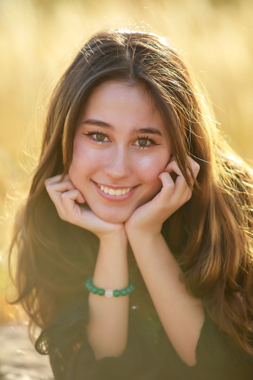 Portrait photo of someone with long brown hair resting their chin on their hands while smiling in warm golden sunlight outdoors.