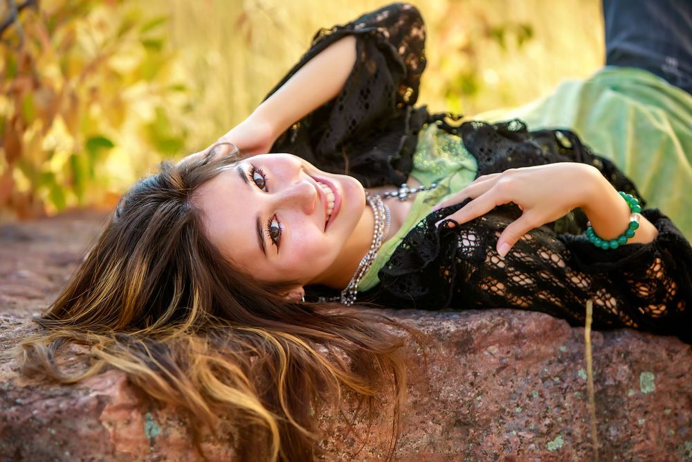 A person with long blonde hair lies on mossy rocks while wearing black clothing as autumn leaves scatter around them.