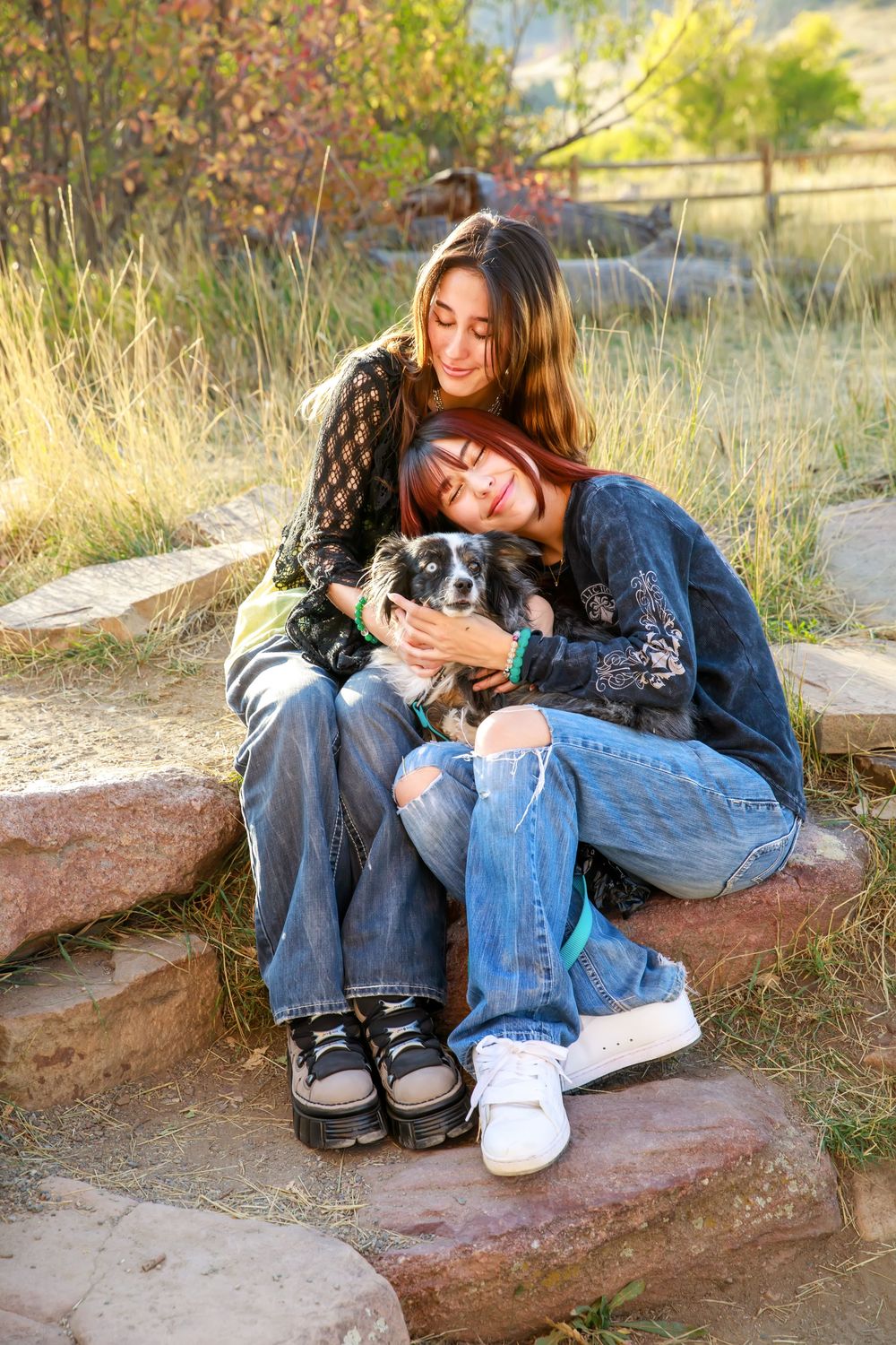 Two friends sitting together on stone steps outdoors with a small dog, enjoying a sunny autumn day in nature.