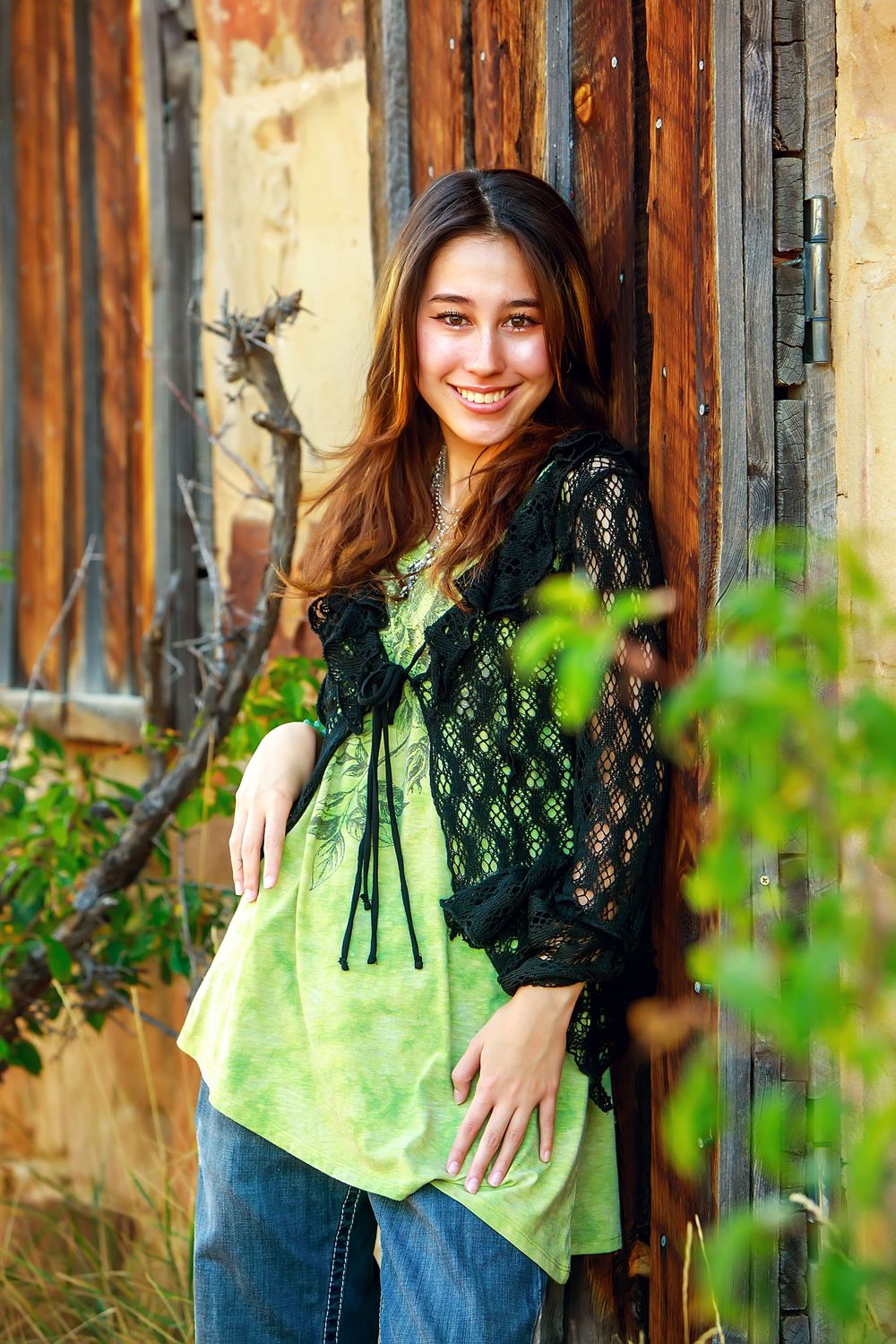 A casual portrait against a rustic wooden door wearing a lime green top with black lace overlay and blue jeans.