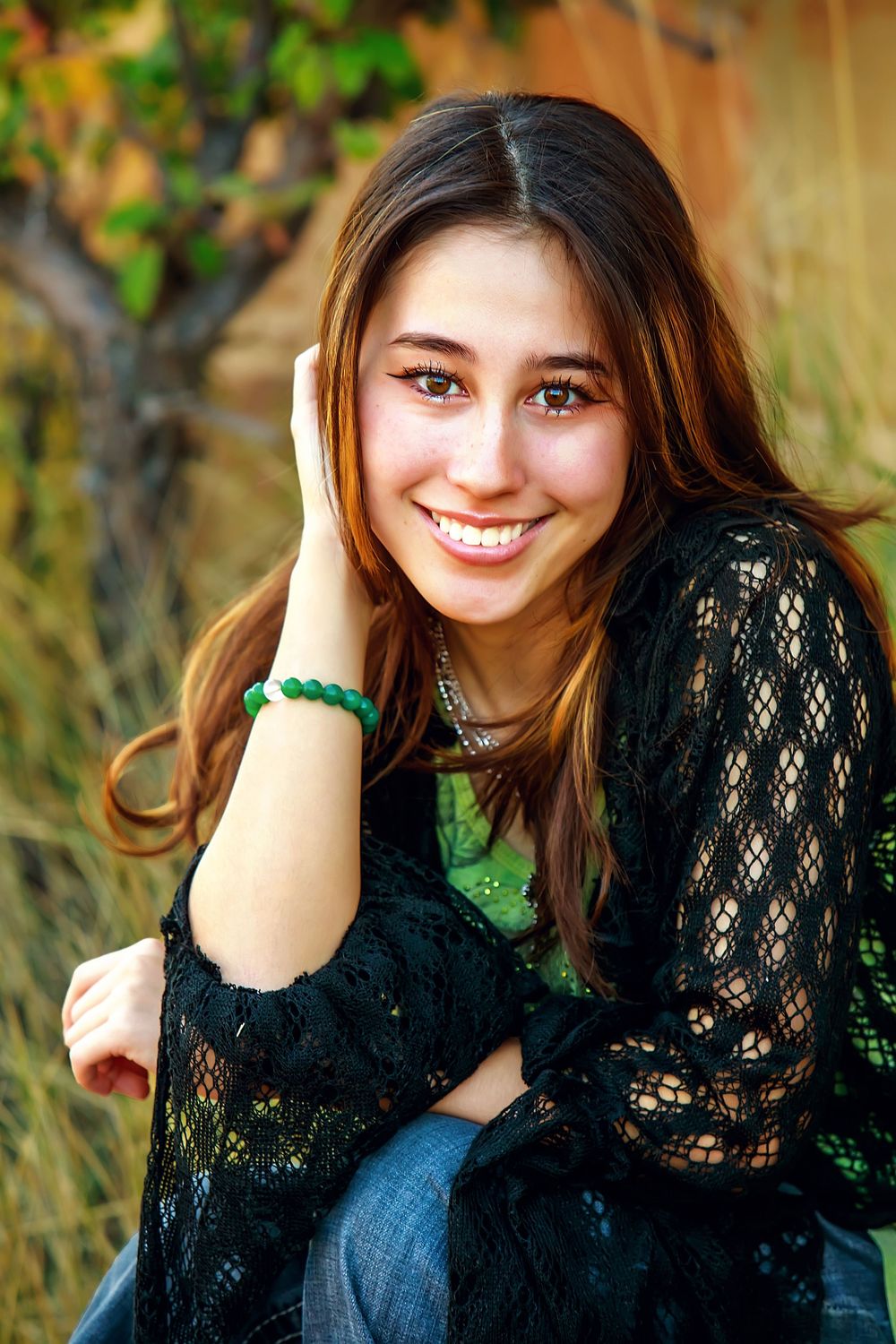 Portrait taken outdoors featuring a black lace top, green bracelet, and jeans against a natural blurred background.
