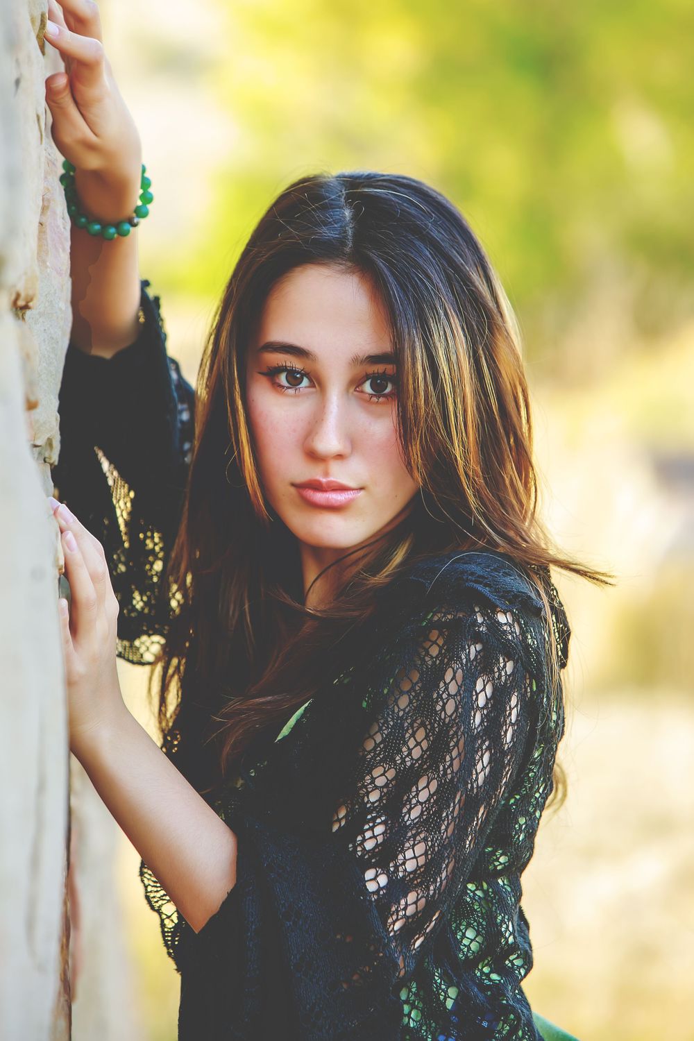 Young woman in black lace top poses against light wall outdoors with soft bokeh background and sunlit effect.